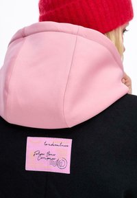 Osoba v černém kabátu s růžovou kapucí a červenou pletenou čepicí, ukazující růžovou nášivku na zádech s nápisem "Následuj svůj kompas za dobrodružstvím."