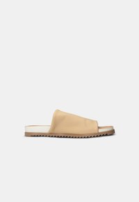 Beige leather slide sandal with an open toe, soft upper design, and a contrasting brown textured rubber sole. Sleek and minimalistic shape.