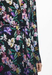 Blumenkleid mit schwarzem Hintergrund, lebhaften, mehrfarbigen Blumen, einer Knopfleiste vorne und einer Bindegürtel-Akzent. Lange Ärmel.