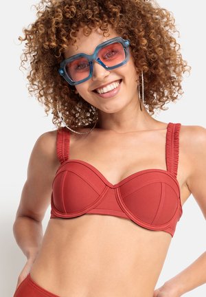 Červený bikini top s drôtikmi, so štruktúrovanými, volánikovými popruhmi. Hladká látka obsahuje detaily švov. Modelka nosí veľké slnečné okuliare s modrým nádychom.
