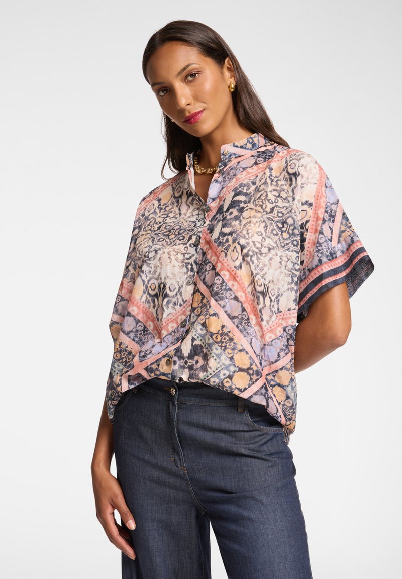 Blusa stampata a maniche corte in morbido tessuto, caratterizzata da una combinazione di colori pastello e motivi geometrici, con colletto e dettagli di bottoni.