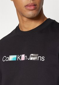 Camiseta negra de manga corta con el texto "Calvin Klein Jeans". Presenta rectángulos de colores en azul, rosa y negro, destacando un diseño gráfico.