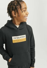 Sudadera negra con capucha que presenta un bolsillo canguro en la parte frontal y un parche rectangular de logo en blanco y naranja con el texto "JACK & JONES ORIGINALS".
