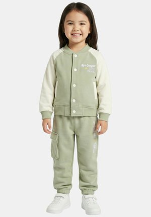 Jeune fille souriante portant une veste vert clair et crème avec un pantalon cargo assorti et des baskets blanches, debout devant un fond blanc.