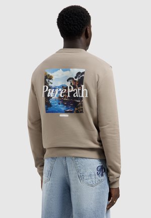 Man wearing beige sweatshirt with a colorful coastal landscape and "Pure Path" text on the back, paired with light blue jeans.