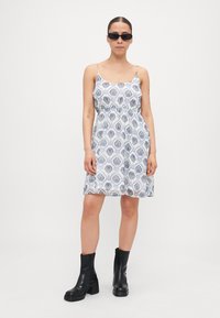 Rochie albă, cu un model floral albastru, cu bretele subțiri și talie strânsă, purtată cu botine negre până la glezne și ochelari de soare închiși la culoare.
