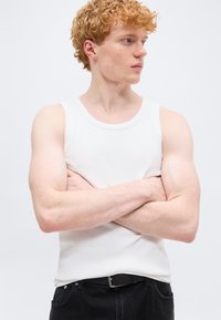 Νεαρός με κόκκινα μαλλιά, φορόντας λευκό αμάνικο πουκάμισο και μαύρο παντελόνι, στέκεται με τα χέρια σταυρωμένα, κοιτάζοντας προς τα δεξιά του μπροστά σε απλό φόντο.