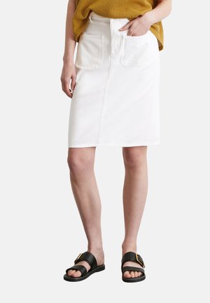 Jupe en denim blanche avec une coupe droite, deux poches avant et une couture centrale. Portée avec un haut en tricot moutarde et des sandales noires.