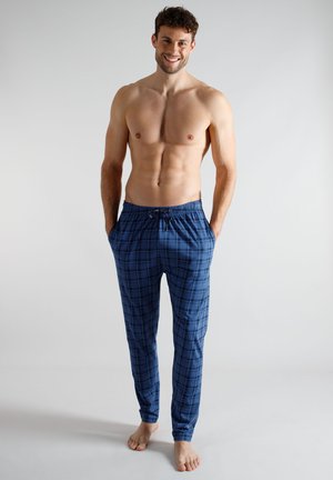 Shirtless man smiling, standing barefoot with hands in pockets, wearing blue plaid pajama pants against plain light background.