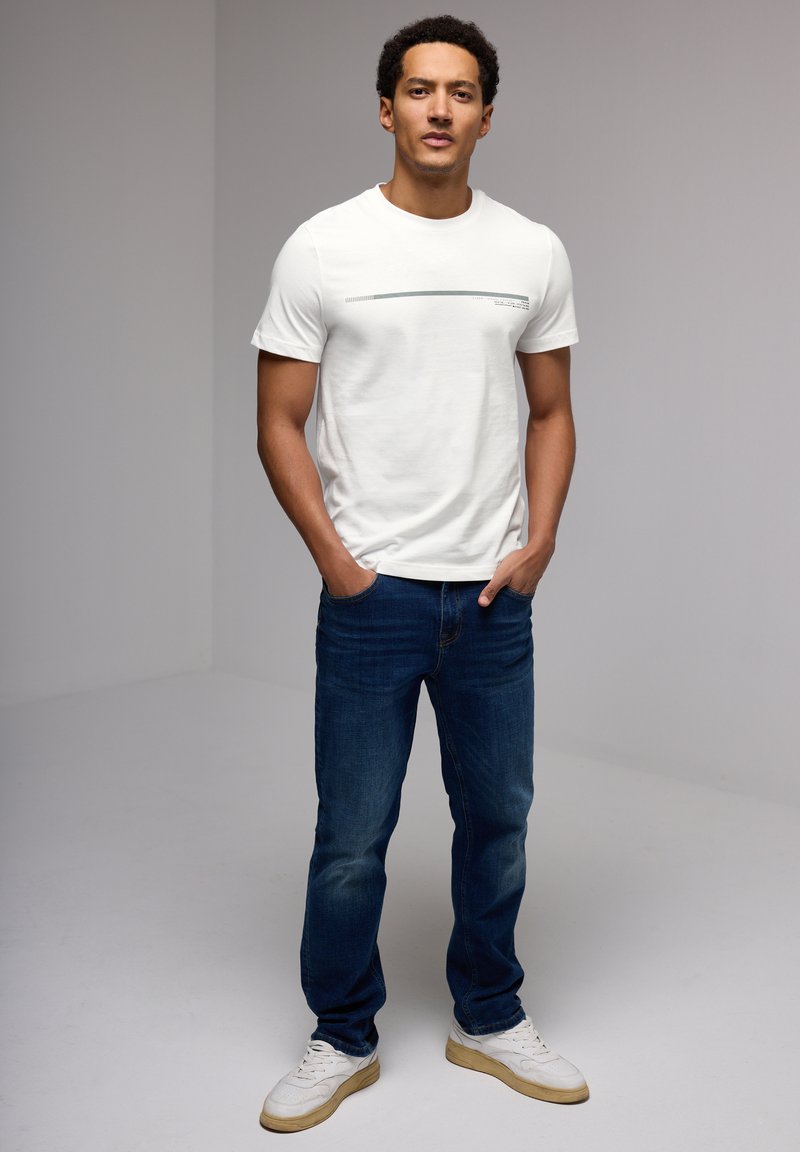 White short-sleeve t-shirt featuring a gray graphic, paired with dark blue jeans and white sneakers with a beige sole.