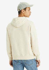 Light beige hoodie with a kangaroo pocket and adjustable hood. Worn with blue jeans; features a gray cap with a buckle closure.