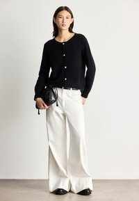 Juoda megzta kardiganas su apvaliu kaklu ir penkiomis mygtukų sagtimis, derinamas su baltais plačiais kelnais ir maža juoda raištelių rankine.