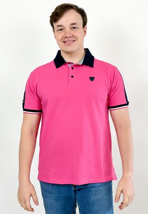 Jeune homme souriant, portant un polo rose vif avec un col noir et un jean bleu, debout devant un fond blanc.