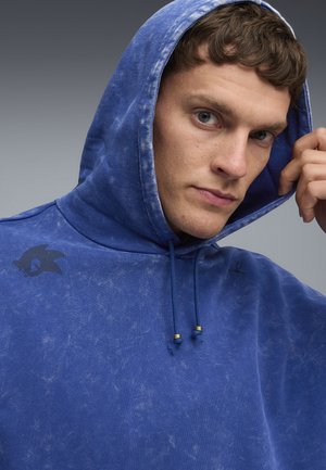 Blå hættetrøje med en slidteknik, der har et mørkt logo på venstre skulder og en snoretræk med guld accenter.