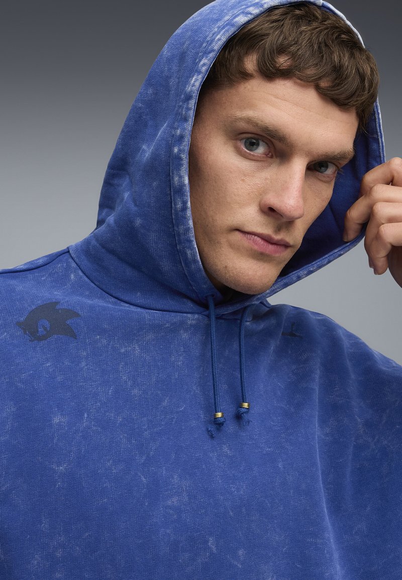 Blå huvtröja med en sliten textur, med en mörk logotyp på vänster axel och en dragsko med guld-accenter.