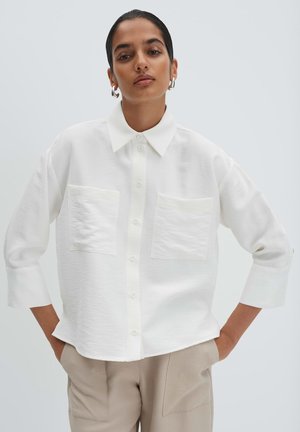 Frau mit dunklen Haaren, die silberne Creolen, ein weißes Hemd mit Knöpfen und zwei Brusttaschen sowie beige Hosen trägt, steht mit den Händen in den Taschen.