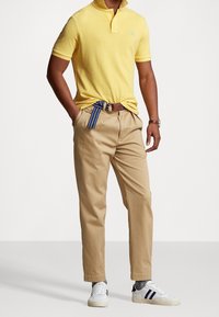 Man i gul polo skjorta, beige byxor med blårandigt bälte, grå strumpor, vita sneakers med marinblå ränder och ett brunt armbandsur.