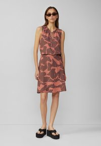 Robe sans manches dans des teintes de rose et de marron avec un col à volants et un motif abstrait. Associée à des sandales à plateforme noires.