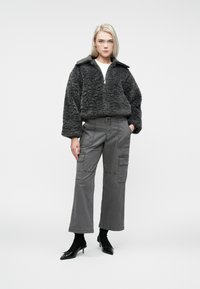 Kvinde iført en mørkegrå, fnugget zip-up jakke, grå cargo bukser, sorte sokker og sorte spidse stiletter, som står med den ene hånd i lommen.