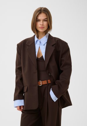 Vrouw met schouderlang haar die een oversized donkerbruin pak draagt, een lichtblauw overhemd met kraag eronder en een bruine riem tegen een grijze achtergrond.