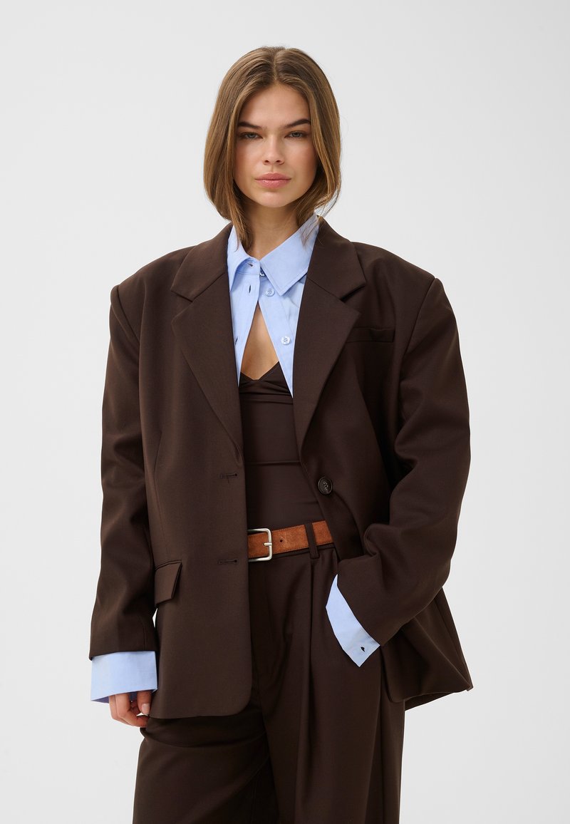 Femme aux cheveux mi-longs portant un costume marron foncé oversize, une chemise bleu clair col boutonné en dessous et une ceinture marron sur fond gris.