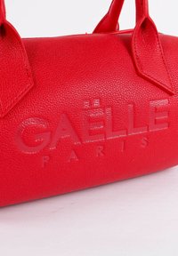 Borsa a mano in pelle rossa con superficie effetto texture e nome del marchio "GAELLE PARIS" in rilievo. Il design include manici corti e una forma compatta.