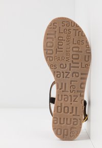Brown rubber sole with textured pattern featuring various logos. Black strap design with a small gold accent on the side.