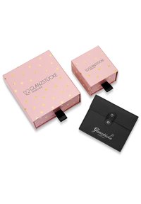 Deux boîtes : une grande rose avec des pois dorés, une plus petite rose, toutes deux avec des languettes noires. Un porte-cartes noir avec logo et fermeture à pression.