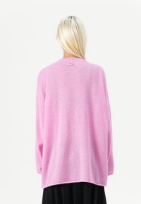 Roza oversize pulover iz mehkega materiala, z okroglo izrezom in majhno etiketo na hrbtni strani z imenom blagovne znamke.