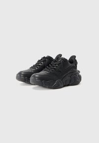 Sorte chunky sneakers med teksturerte tykke såler, snøring og subtil logo-detalj på siden, plassert på en enkel hvit bakgrunn.