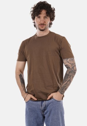 Uomo con capelli ricci e tatuaggi su tutto il braccio, indossa una maglietta marrone e jeans blu, con le mani nelle tasche, in piedi contro uno sfondo neutro.