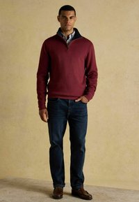 Jonge man staat in een bordeaux kwart-rits trui over een geruit shirt, donkere spijkerbroek en bruin leren instapschoenen, tegen een beige achtergrond.