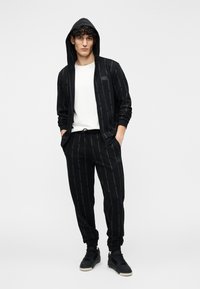 Sudadera negra con cremallera y patrón repetido de logotipos, combinada con pantalones deportivos a juego, ambos hechos de tela suave. Camiseta blanca debajo.