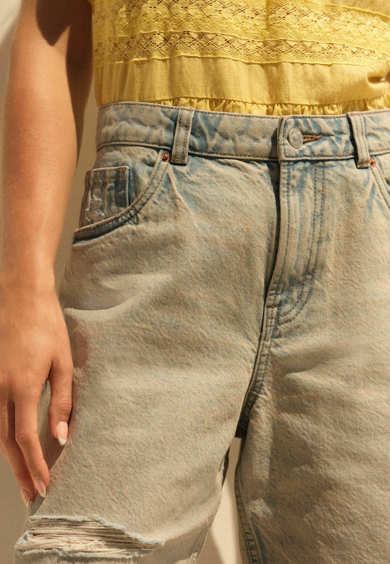 Pantalones cortos de denim azul claro con un acabado desgastado, detalles rasgados, cinco bolsillos y costuras visibles. Combinados con una blusa amarilla de textura.