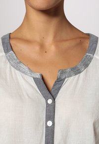 Camicia bianca con colletto grigio e finiture metalliche. I bottoni sono bianchi e il tessuto sembra leggero e strutturato.