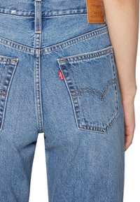 Svetle modré džíny z denimu s vysokým pasem, mají dvě zadní kapsy a oranžové švy. Obsahují hnědou koženou značkovou visačku.