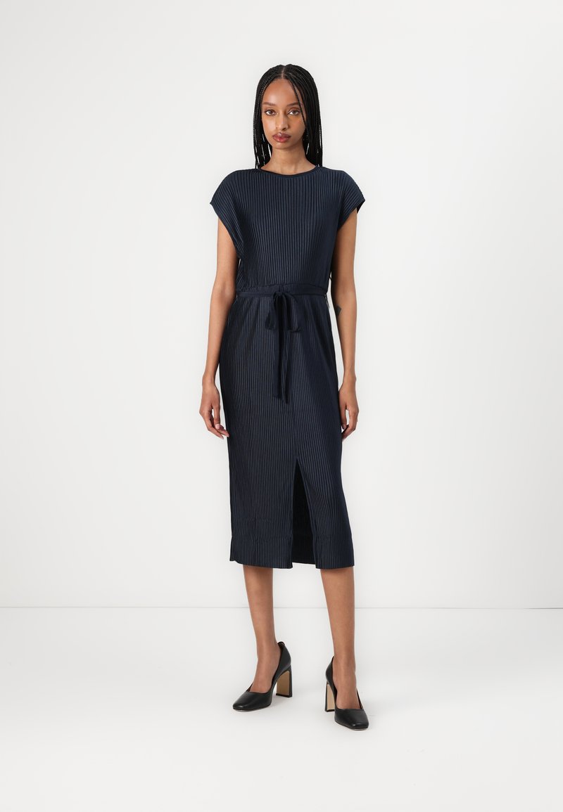 Abito midi a righe navy con maniche a cappuccio, vita con cintura e spacco laterale, abbinato a tacchi neri. Tessuto liscio con linee verticali.