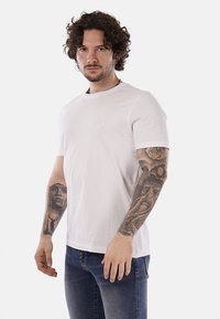 T-shirt bianca in cotone con collo alla marinara e maniche corte, caratterizzata da un sottile logo impresso sul petto. Indossata con jeans blu.