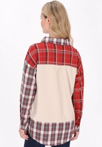 La camicia presenta una combinazione di motivi scozzesi nei colori rosso, bianco e grigio, con un retro in velluto a coste beige e un orlo curvo. Chiusura a pulsante ai polsi visibile.