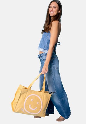 Woman in denim outfit smiling, holding large yellow tote bag with pink smiley face, standing against white background.