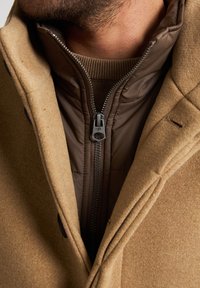 Cappotto beige con un esterno in lana testurizzata e una giacca interna imbottita marrone. Presenta una cerniera visibile e bottoni, con un primo piano dell'area del colletto.
