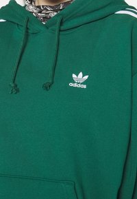 Zielona bluza z kapturem Adidas z białym logo trefoil na piersi i sznurkami zwisającymi z kaptura. Osoba nosi pod spodem wzorzysty apaszkę na szyi.