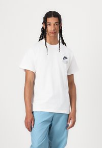 Balta medvilninė marškinėliai su mėlynu Nike logotipu kairėje krūtinės pusėje, trumpomis rankovėmis ir laisvu siluetu, derinami su šviesiai mėlynomis kelnėmis.