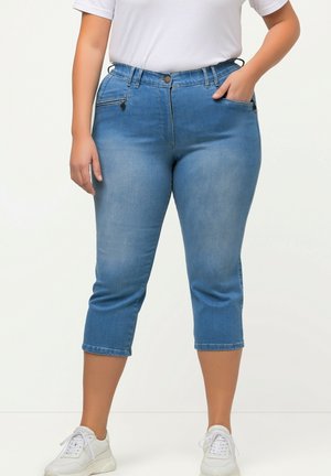 Jean capri en denim bleu clair avec fermeture à bouton, deux poches avant et une légère décoloration. Assorti à des baskets blanches et un t-shirt blanc.