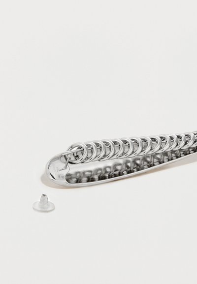 Μεταλλική παραμάνα ασφαλείας με λαμπερό ασημί φινίρισμα, που διαθέτει μια σειρά από διασυνδεδεμένους κρίκους, και ένα ξεχωριστό διάφανο πλαστικό στοπ δίπλα της.