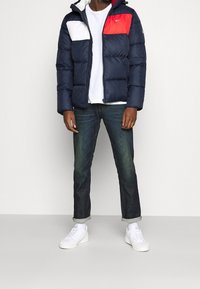 Granatowa kurtka puchowa z białymi i czerwonymi akcentami, pikowany wzór, szare dżinsy i białe sneakersy. Model z kapturem, zapinany na zamek.