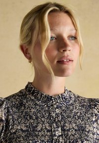 Ziedu raksta blūze tumši zilā un krēmkrāsā, ar smockētu tekstūru, augstu apkakli un īsām pūkainām piedurknēm. Redzami zelta riņķa auskari.