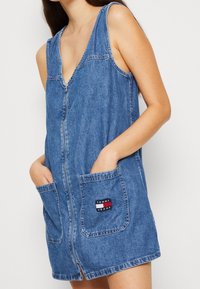 Kobieta w niebieskiej sukience z dżinsu bez rękawów, z zamkiem błyskawicznym z przodu i dwiema kieszeniami, jedna z kieszeni posiada naszywkę z logo Tommy Jeans.