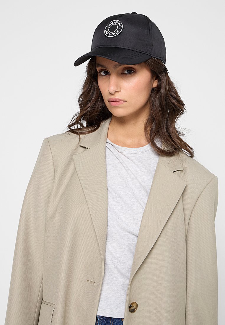 Casquette de baseball noire avec un logo circulaire sur le devant, associée à un blazer beige et un t-shirt gris. Tissu lisse avec un design structuré.