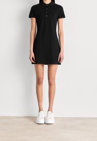 Svart poloklänning med krage, korta ärmar och broderad logotyp. I kombination med vita sneakers på en ljus texturerad yta.